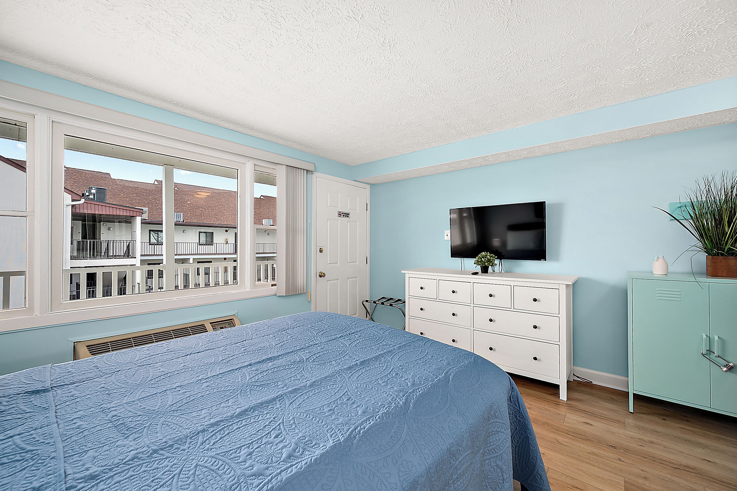 A bedroom with a large bed, a dresser, a television, and a view of a balcony with buildings.