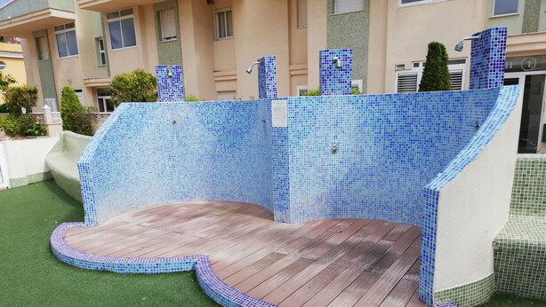 Une piscine aux carreaux de mosaïque bleus, une terrasse en bois et une pelouse artificielle verte.