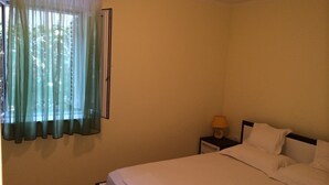 Une chambre à coucher avec un lit, une table de chevet, une lampe et une fenêtre ornée de rideaux légers.