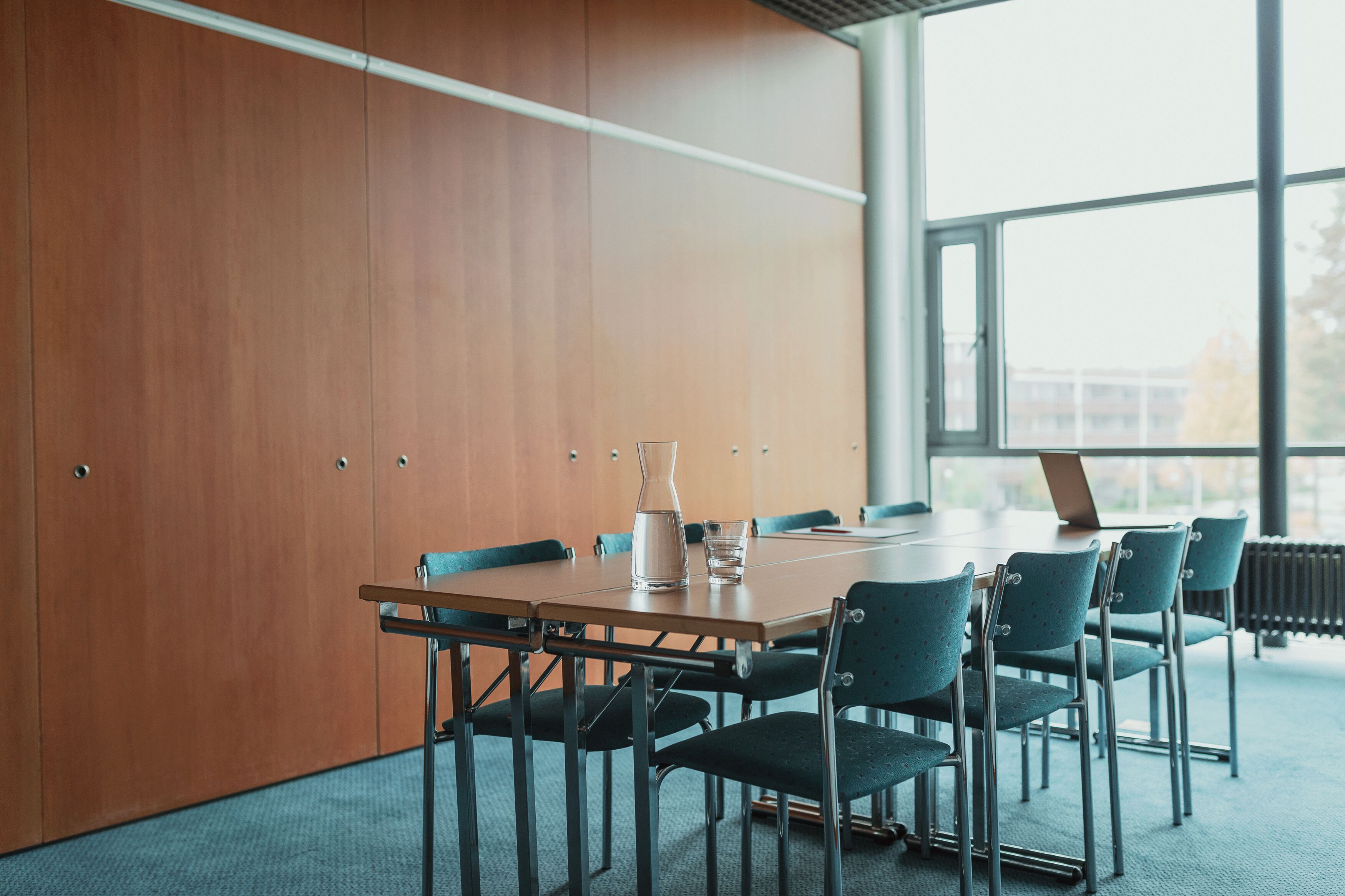 A conference room with a wooden panel wall, a large window, a table with chairs, and a laptop.