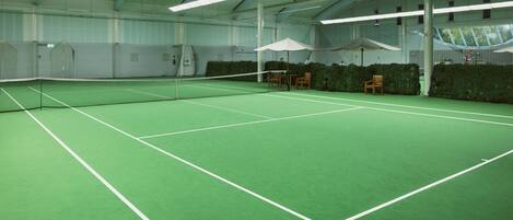Pista de tenis interior con red, suelo verde y zonas de estar con sombrillas.