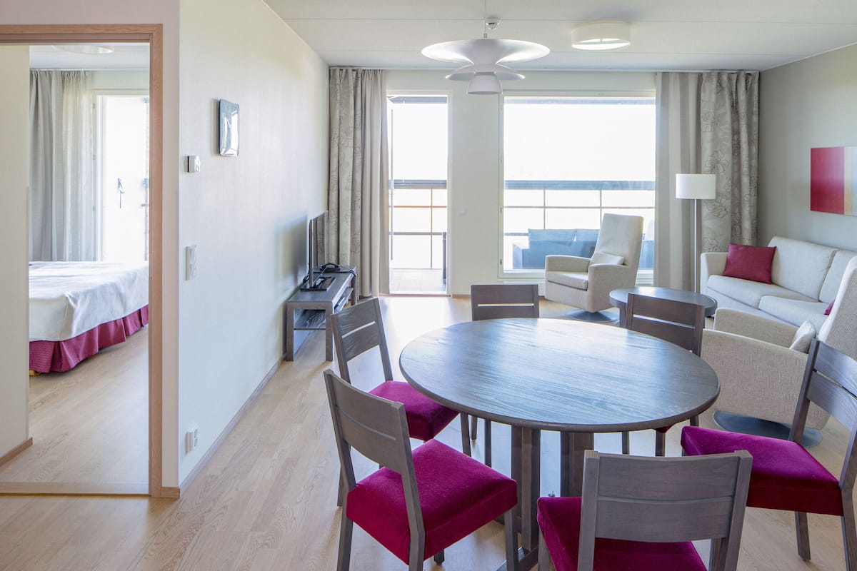 Habitación de hotel con cama, mesa de comedor con sillas, sofá y balcón con cortinas.
