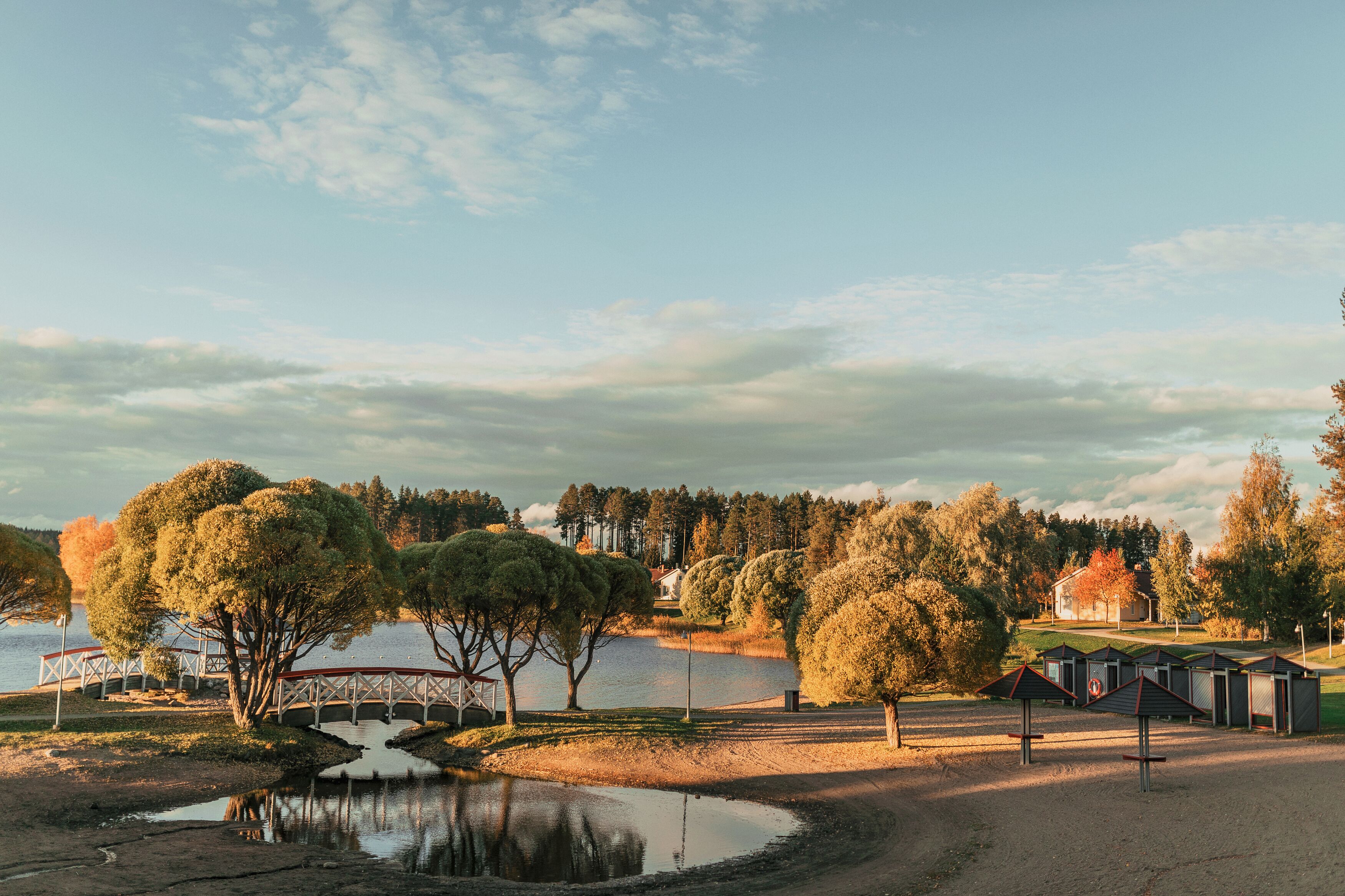Un parque junto al lago con un puente de madera, mesas de picnic y un pequeño edificio.