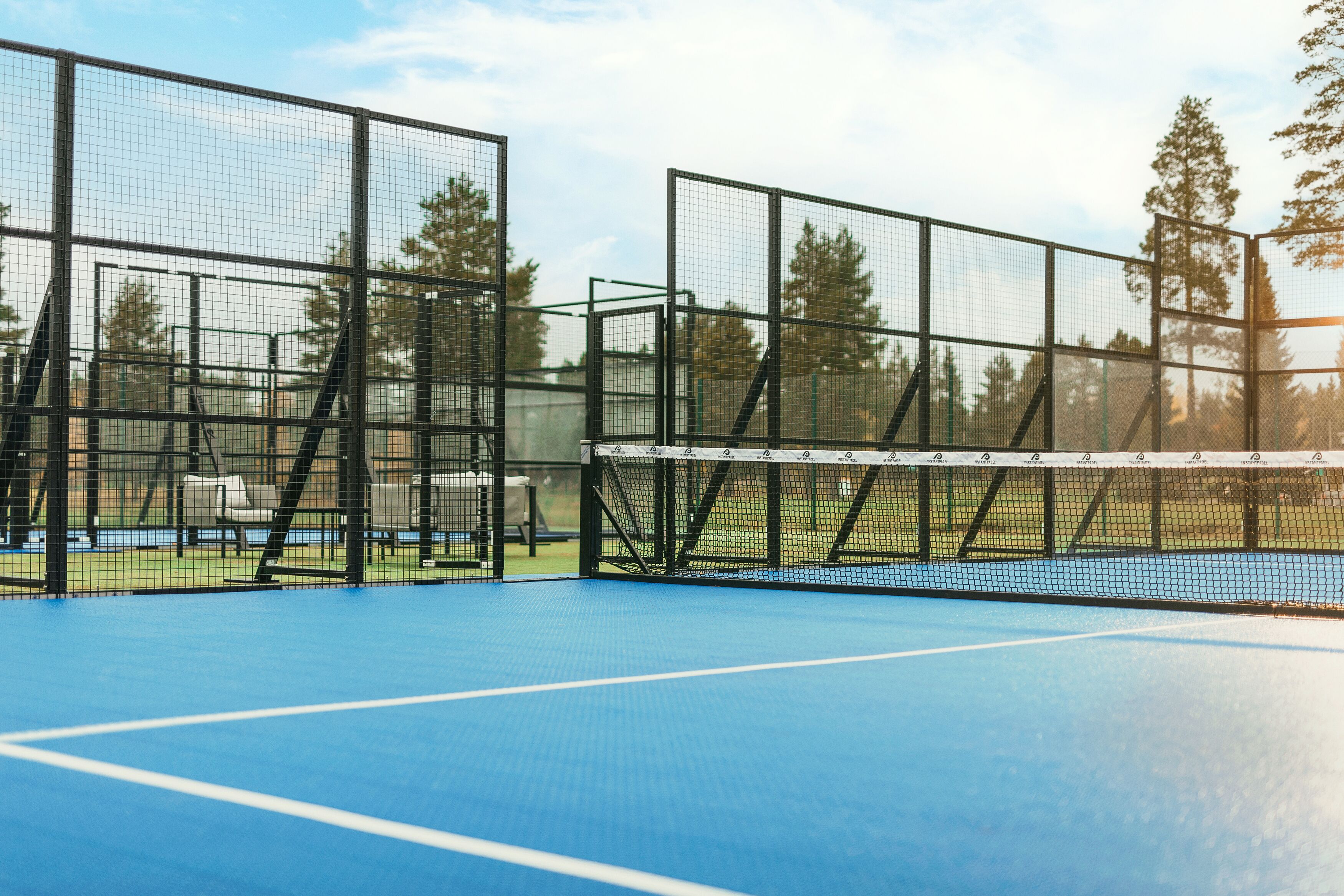A tennis court with a net, surrounded by a metal fence, and trees in the background.