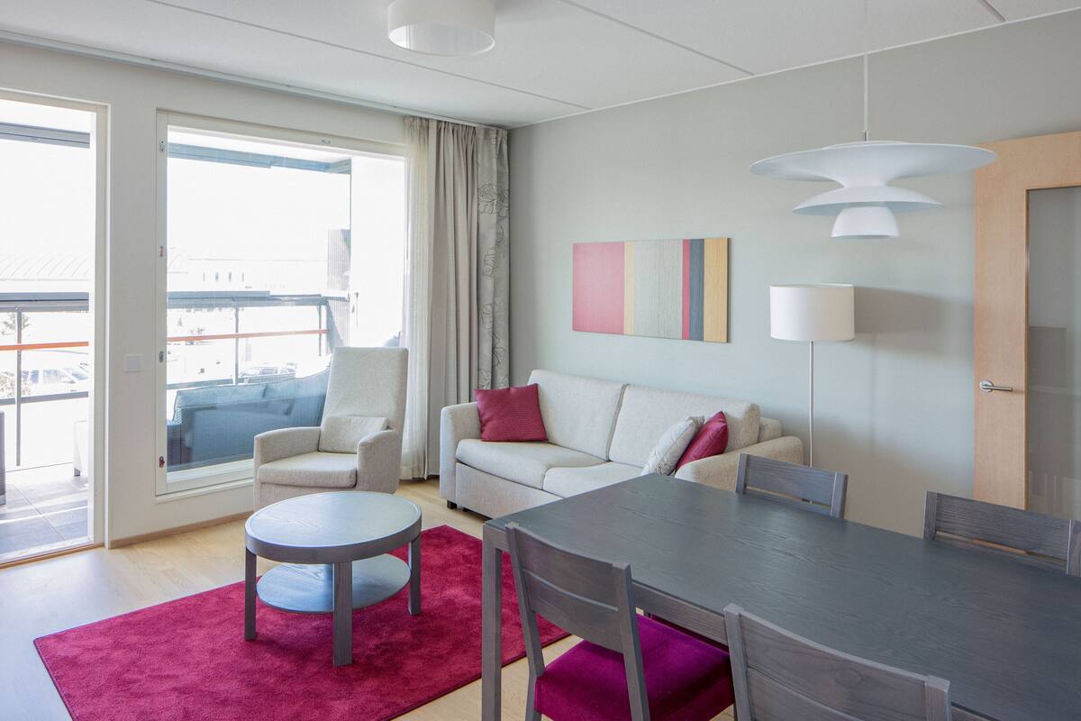 Habitación de hotel moderna con sofá, sillón, mesa de comedor y un ventanal con vistas a la ciudad.
