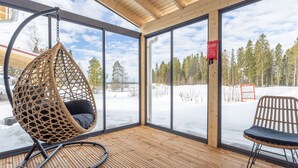 Une terrasse en bois avec un fauteuil suspendu, une chaise et une vue sur un paysage enneigé, visible à travers de grandes fenêtres.