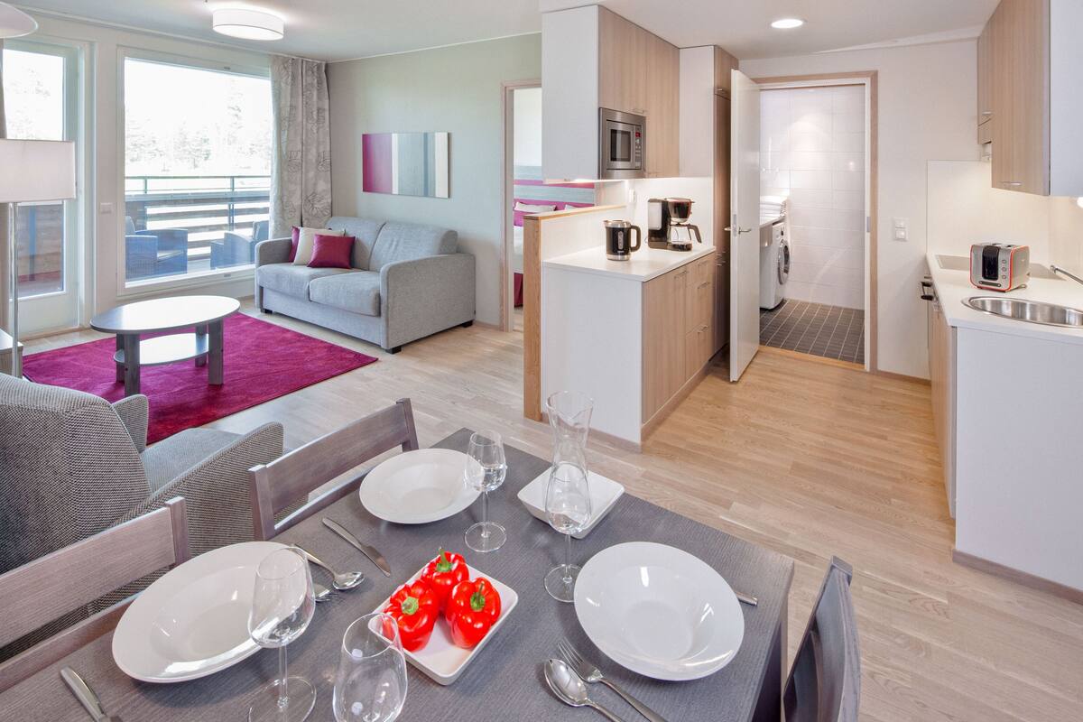 Una sala moderna con un sofá gris, una mesa de comedor para dos personas y una zona de cocina con microondas y cafetera.