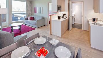 Een moderne woonkamer met een grijze bank, een eettafel voor twee personen en een keuken met een magnetron en koffiezetapparaat.