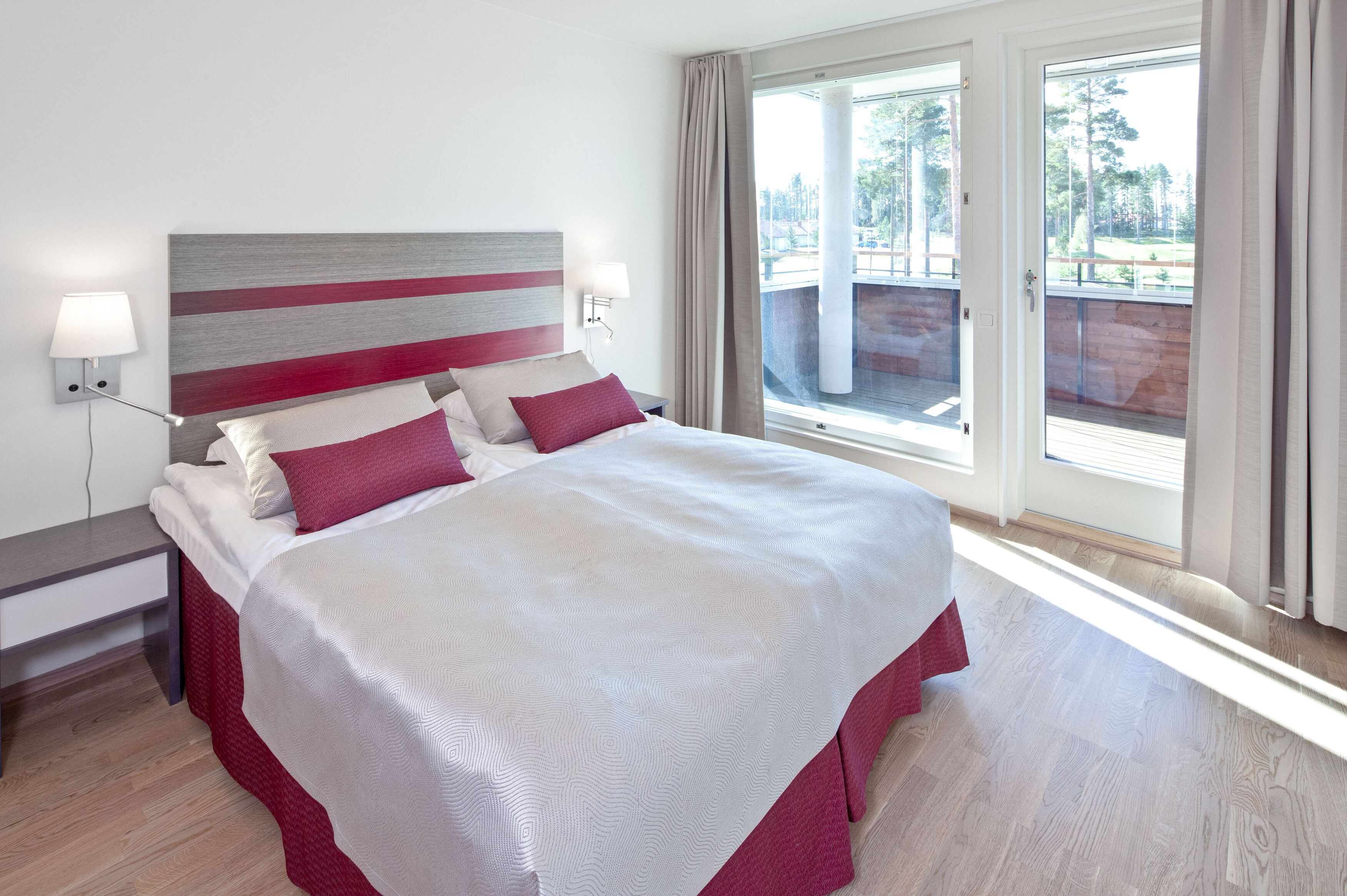 A neatly made bed with a striped headboard, a bedside table with a lamp, and a view of a balcony through a large window.