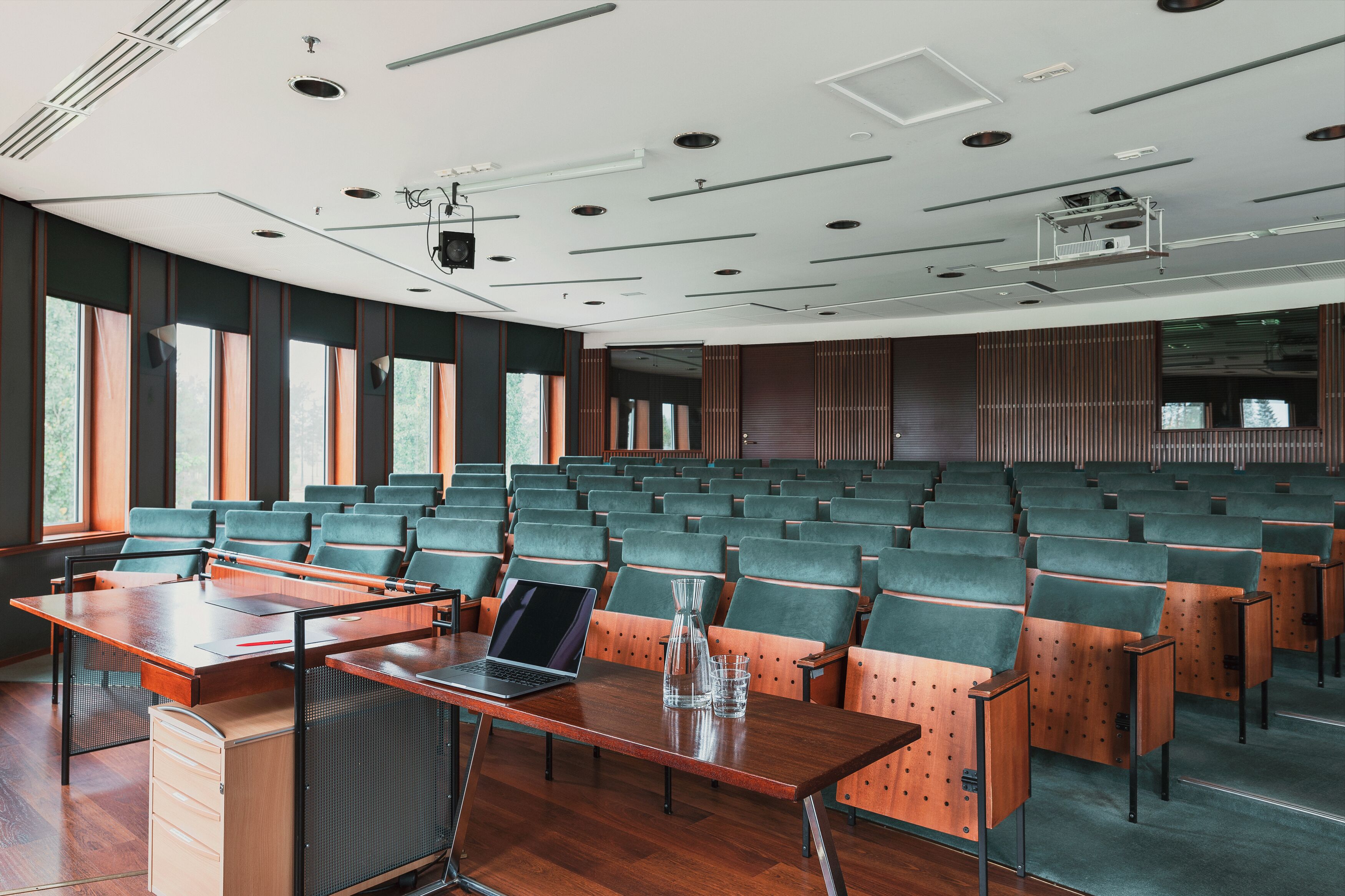 A conference room with rows of green chairs, a wooden table with a laptop, and a projector on the ceiling.