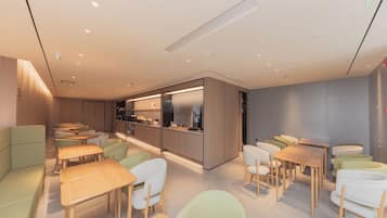A modern dining area with wooden tables and chairs, a kitchen area, and a seating area with cushioned benches.
