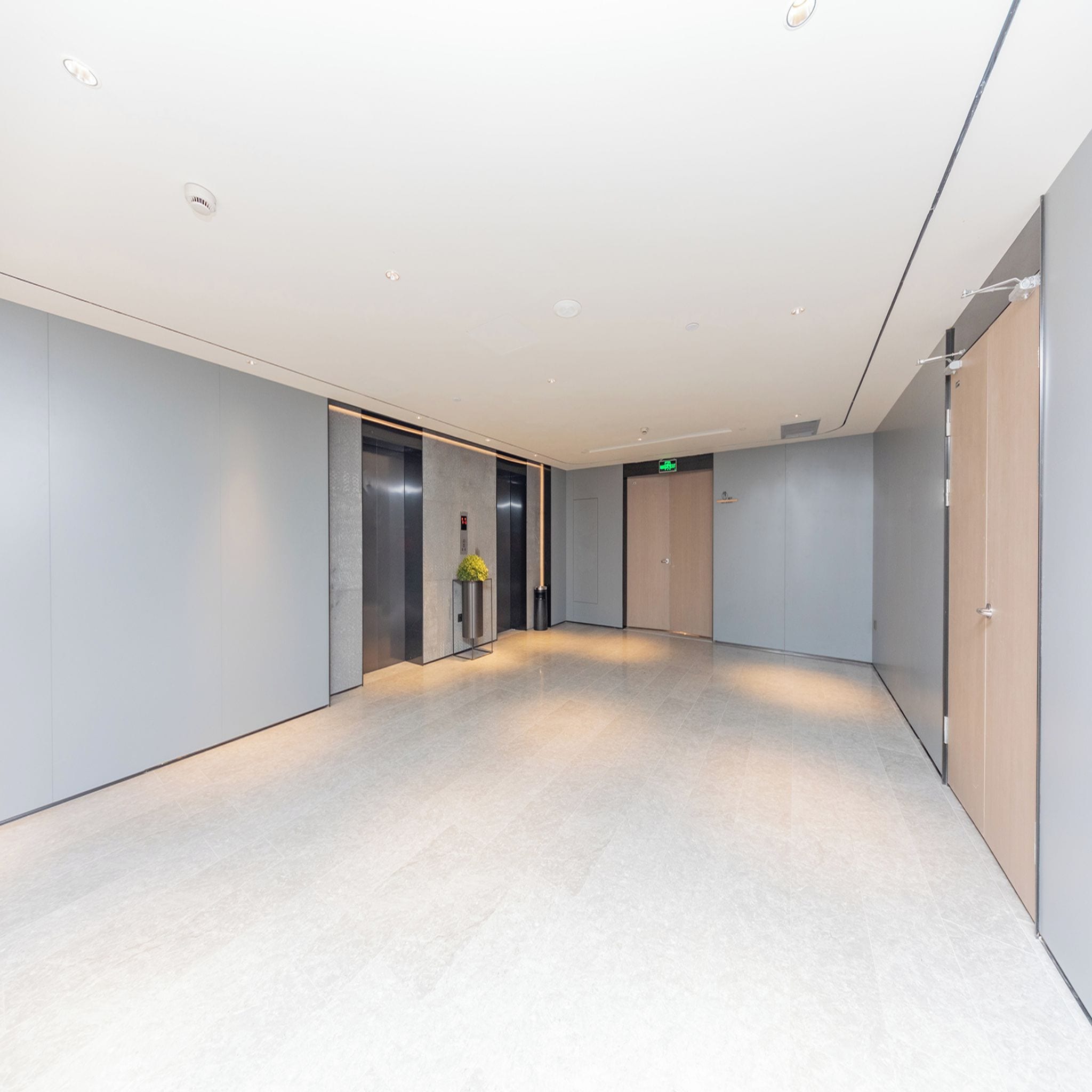 A modern hallway with a light-colored floor, a potted plant, and a green exit sign.