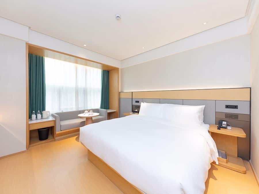 Quarto de hotel moderno com uma cama grande, mesas de cabeceira, uma pequena mesa redonda e uma janela com cortinas.