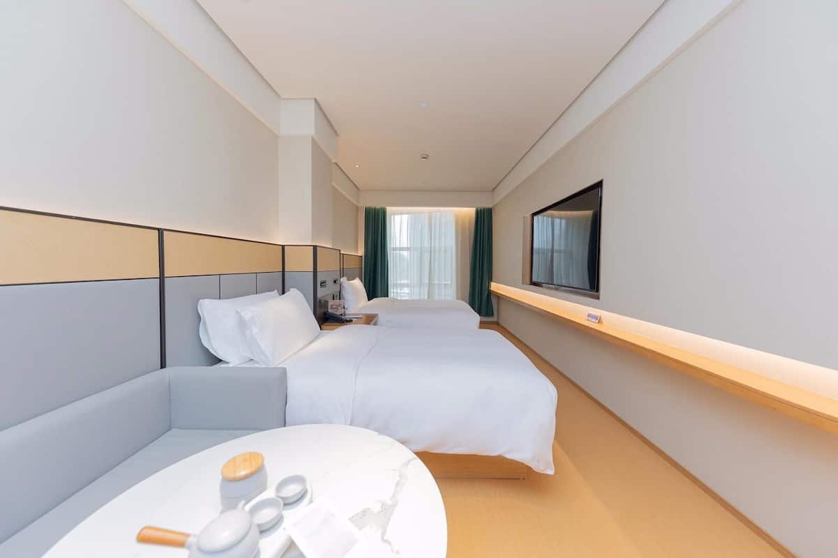 Et hotelværelse med to senge, et tv ophængt på væggen, et lille bord med toiletartikler og udsigt til gangen.