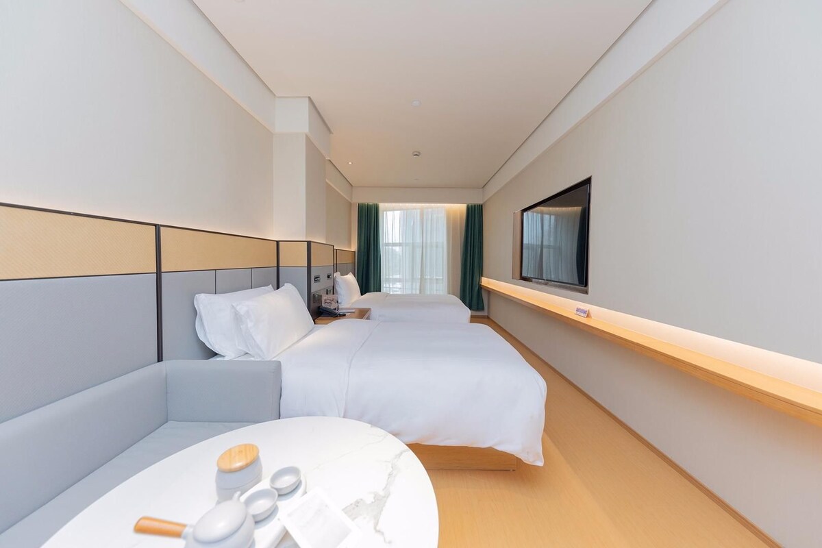 A hotel room with two beds, a TV mounted on the wall, a small table with toiletries, and a view of the hallway.