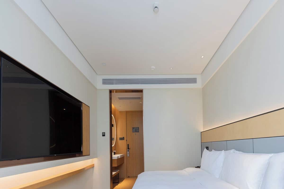 Et moderne hotelværelse med en stor seng, et fladskærms-TV monteret på væggen, et natbord med en lampe og et badeværelse, der kan ses gennem en åben dør.