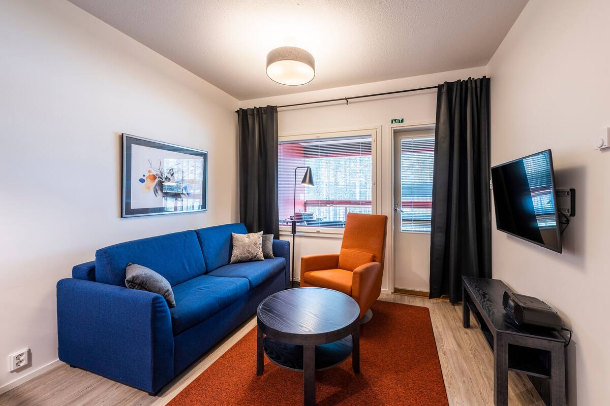 Una sala de estar moderna con un sofá azul, un sillón naranja, una mesa redonda, un televisor de pantalla plana y un cuadro en la pared.