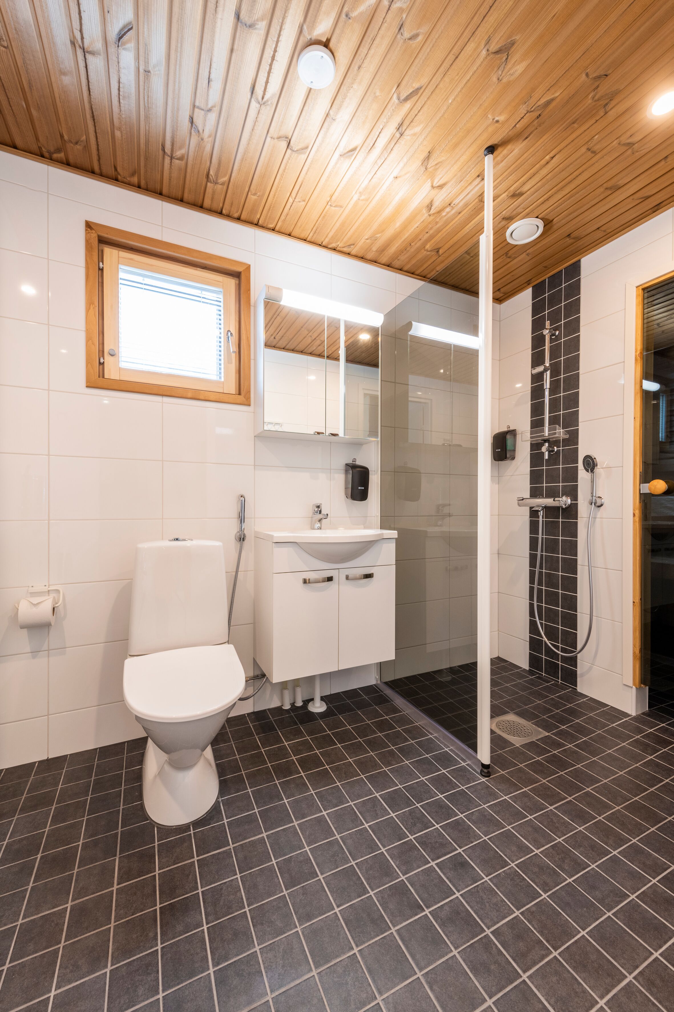 Un baño moderno con techo de madera, inodoro blanco, lavamanos con espejo y zona de ducha.