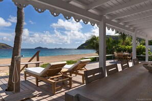 Terraza de madera con sillones y una mesa, con vista a una playa de aguas azules cristalinas y islas al horizonte.