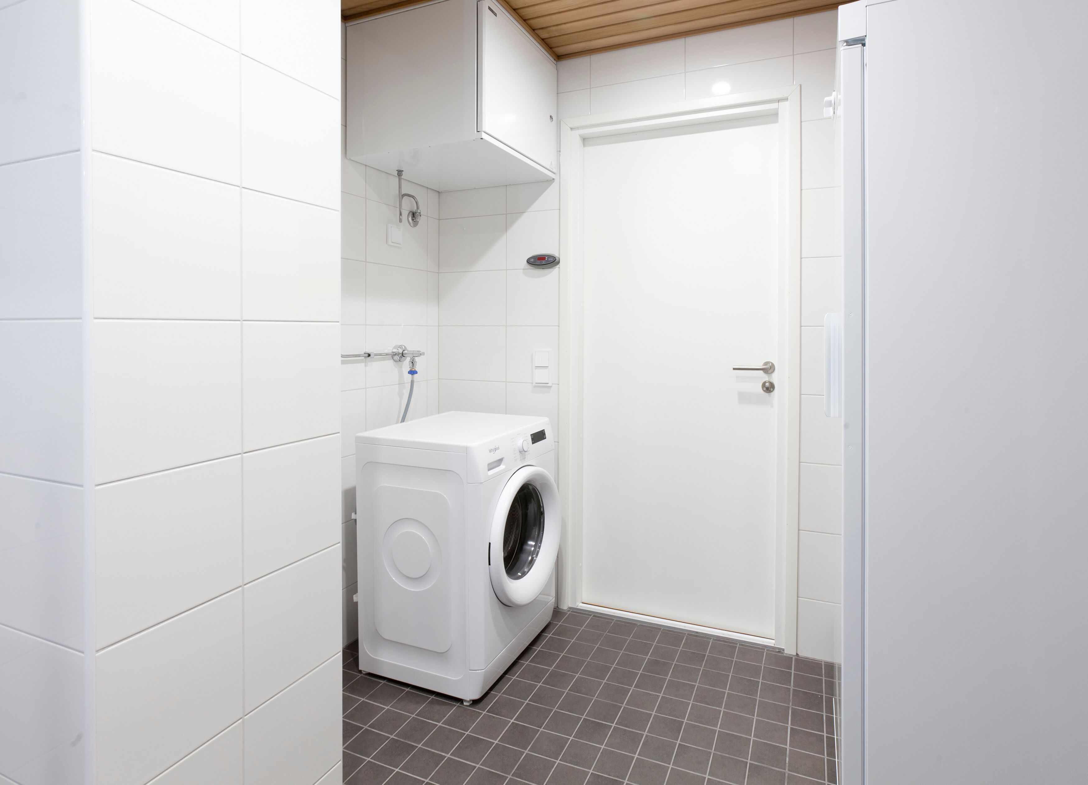 Une salle de bain moderne et compacte, dotée d’un lave-linge et d’un sèche-linge intégrés, de murs recouverts de carreaux blancs et d’un sol carrelé de couleur grise.