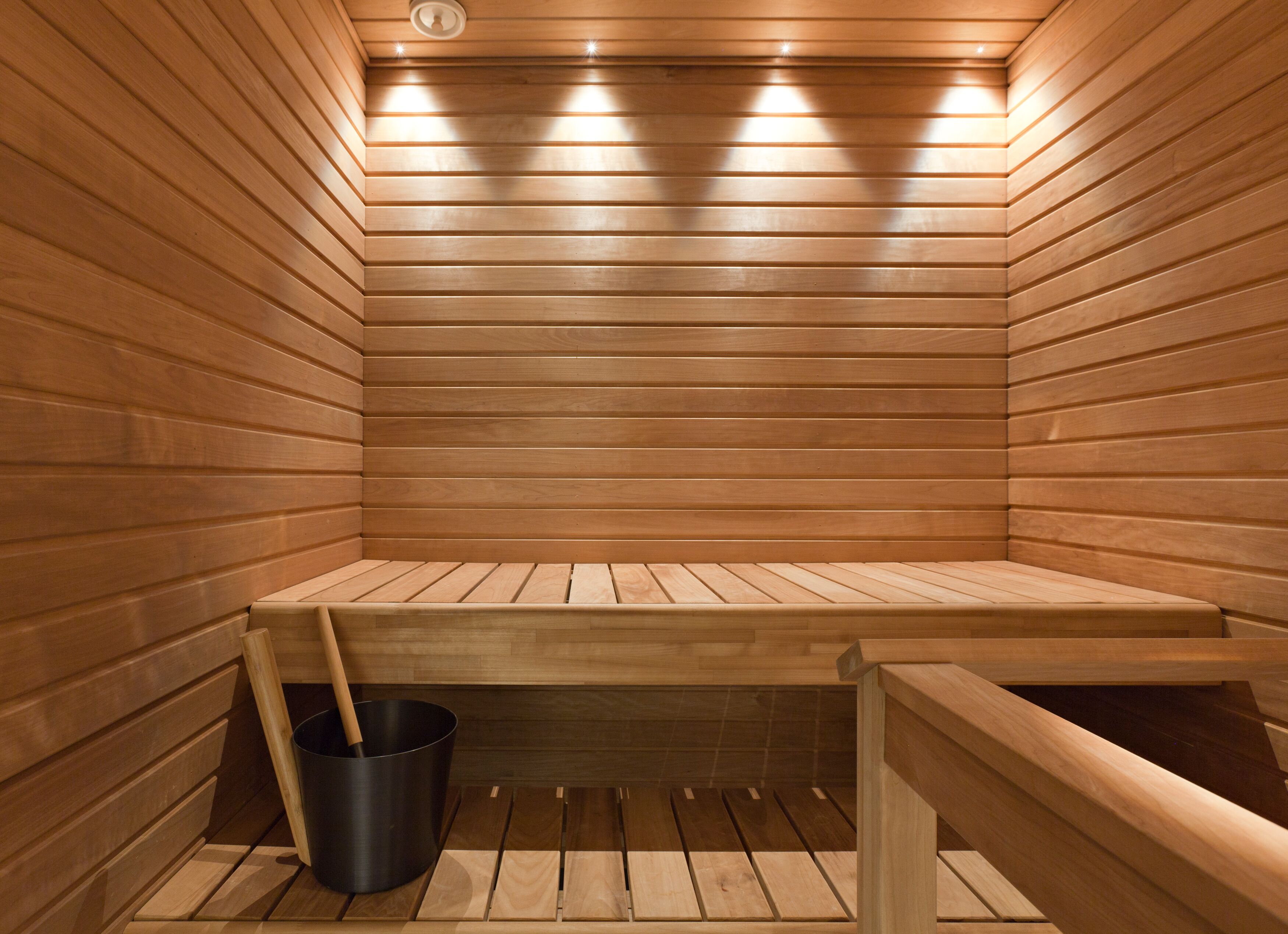 A sauna with wooden walls and benches, a bucket, and a ceiling light fixture.
