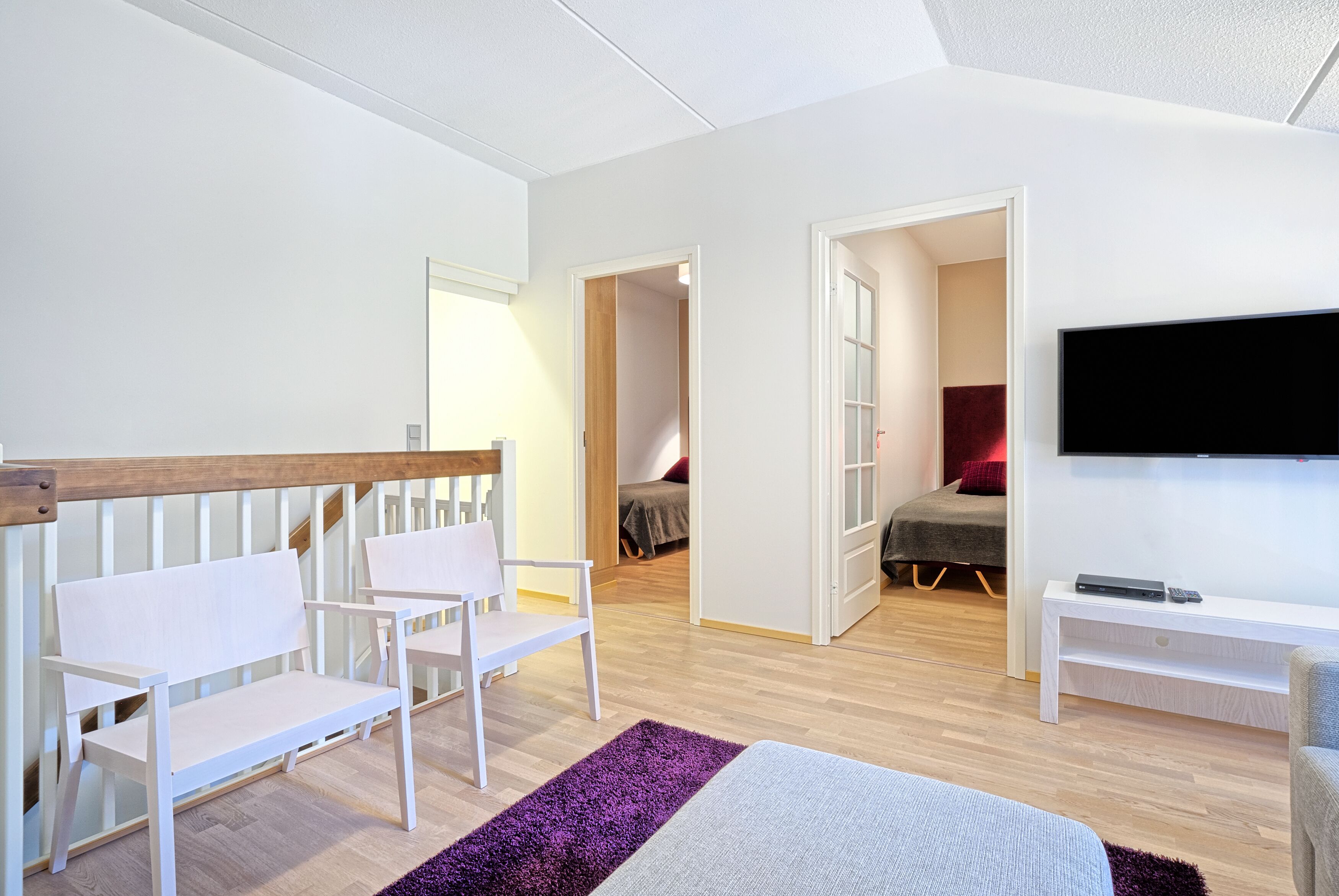 Una habitación de hotel moderna con una cama, dos sillas blancas, un televisor y un banco de madera.