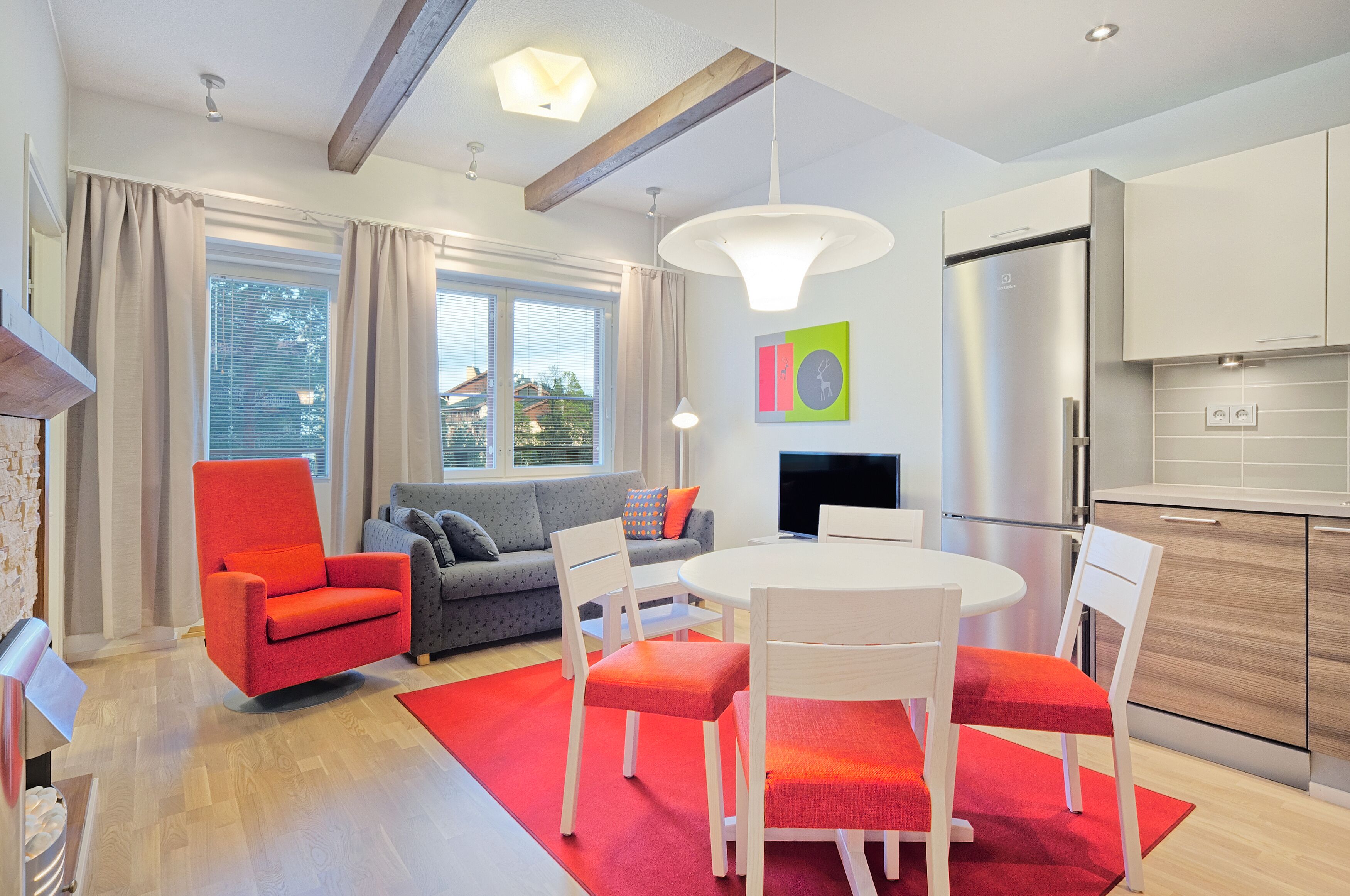 A modern living room with a red armchair, grey sofa, and a dining table with white chairs. There is a kitchen area with a stainless steel refrigerator and wooden cabinets.