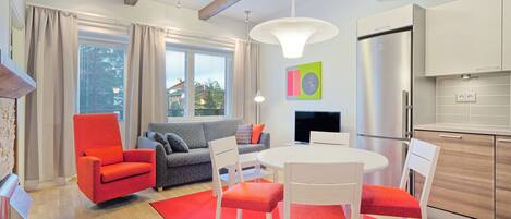 Ein modernes Wohnzimmer mit einem roten Sessel, einem grauen Sofa und einem Esstisch mit weißen Stühlen. Es gibt eine Küchenzeile mit einem Edelstahl-Kühlschrank und Holzschränken.