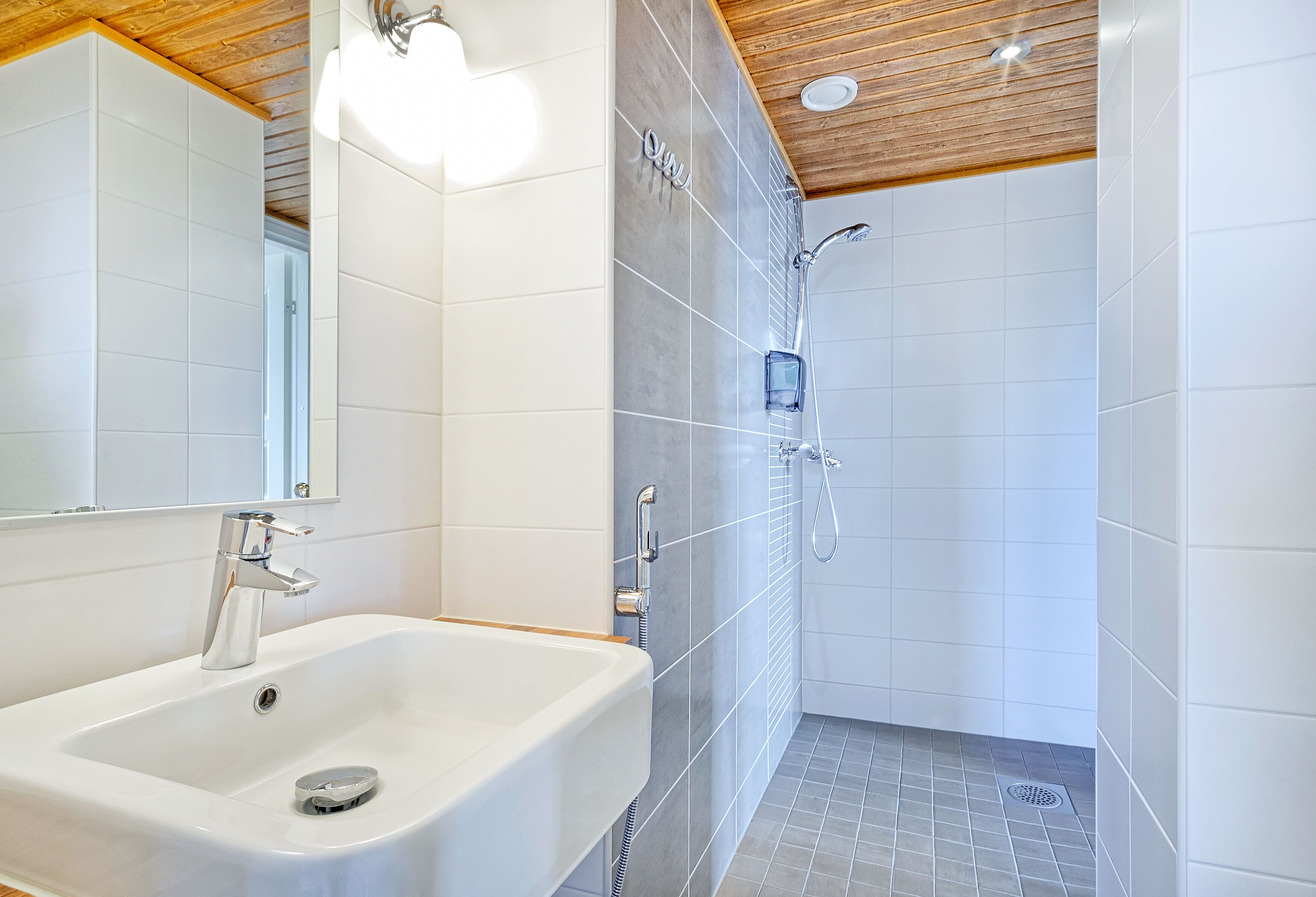 Une salle de bain moderne avec un lavabo blanc, un miroir et un espace douche doté d’un mur et d’un plafond carrelés.