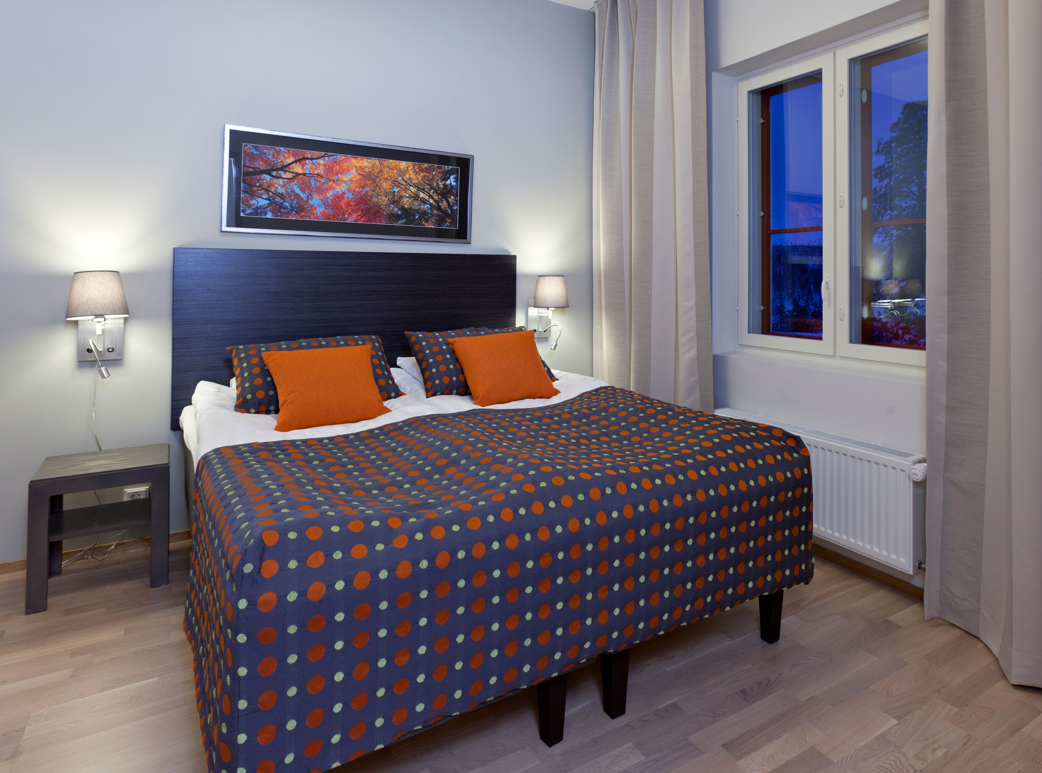 Una cama bien hecha con una colcha de lunares, dos mesitas de noche con lámparas, un cuadro enmarcado en la pared y una ventana con cortinas.