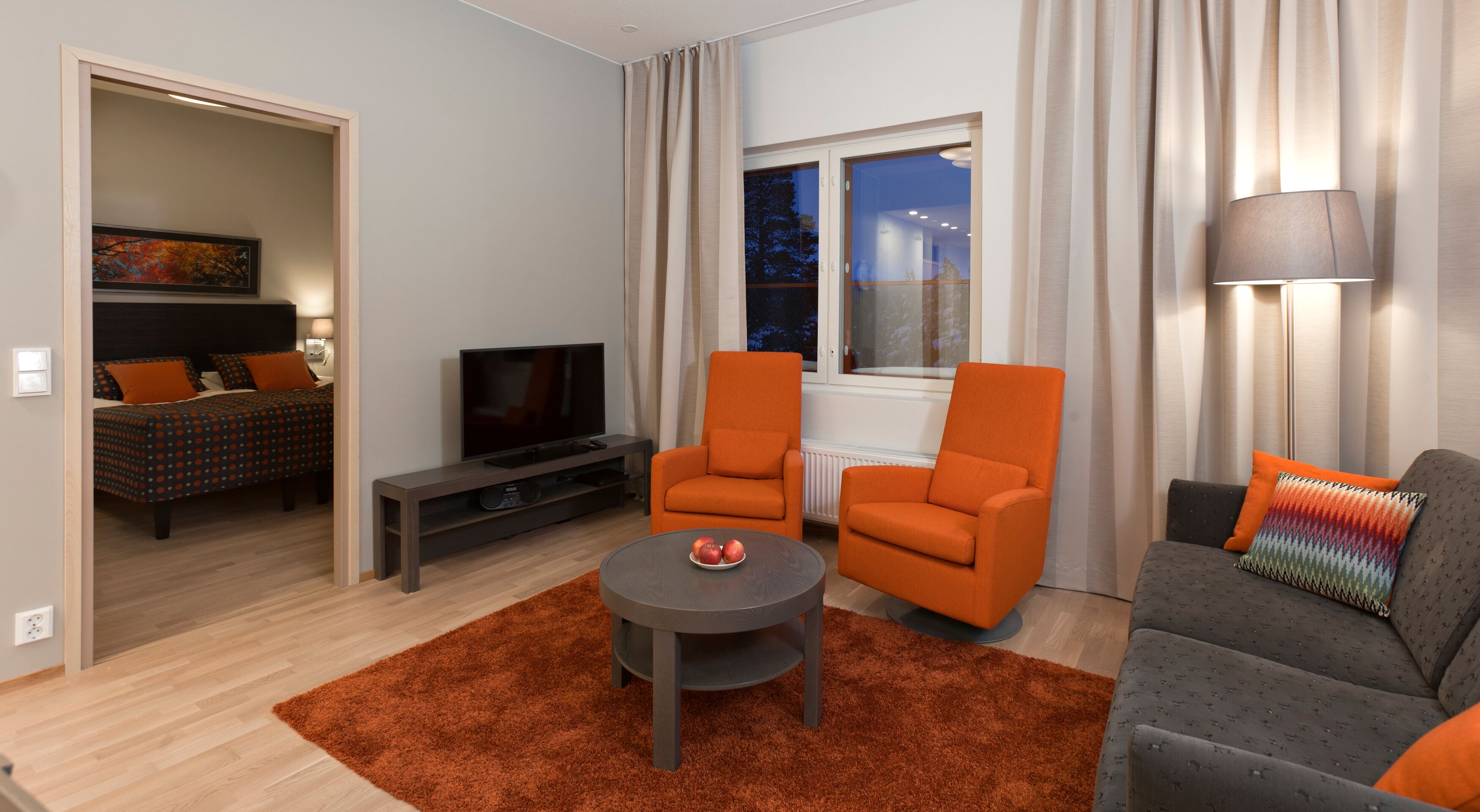 Un salon moderne avec un téléviseur, deux fauteuils orange, un canapé gris et une petite table basse sur laquelle sont posées des pommes.