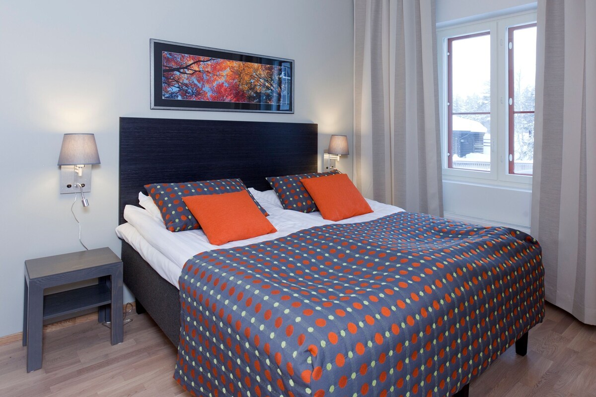 Una habitación de hotel moderna con una cama grande, dos lámparas de noche, un cuadro en la pared y un ventanal con cortinas.