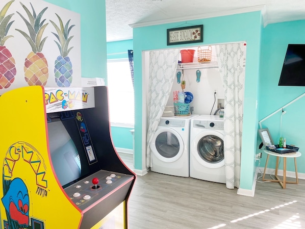 A laundry area with two washing machines, a small table with a chair, and a colorful arcade machine. - Mermaid Dreams ~ Cozy getaway less than 1/4 of a mile from the beach! (Panama City Beach)