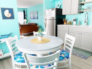 A dining area with a round table, white chairs with blue cushions, a small potted plant, and a kitchen in the background. - Mermaid Dreams ~ Cozy getaway less than 1/4 of a mile from the beach! (Panama City Beach)