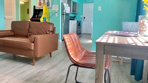 Un salón con un sofá de cuero marrón y una mesa de comedor de madera con una silla. Se ve una cocina al fondo, con un refrigerador y un microondas.