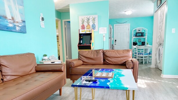 Un salón con un sofá de cuero marrón, una mesa de centro de vidrio con diseño azul y blanco, y una zona de cocina con lavadora y microondas.