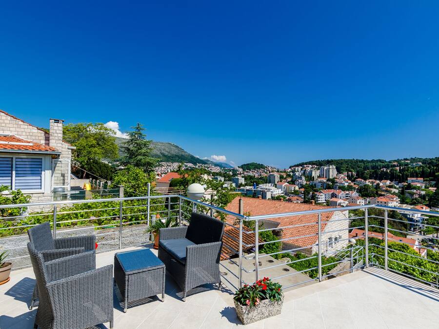 Terraza en la azotea con mobiliario de mimbre y plantas en macetas, con vistas a un paisaje urbano con edificios de tejados rojos y áreas verdes.