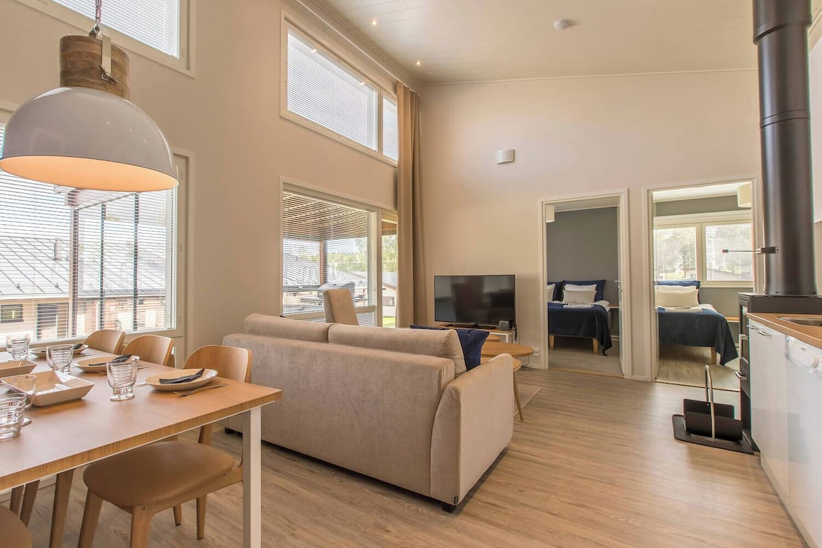 A modern, well-lit living room with a sofa, dining table, and TV. There are large windows and a fireplace.