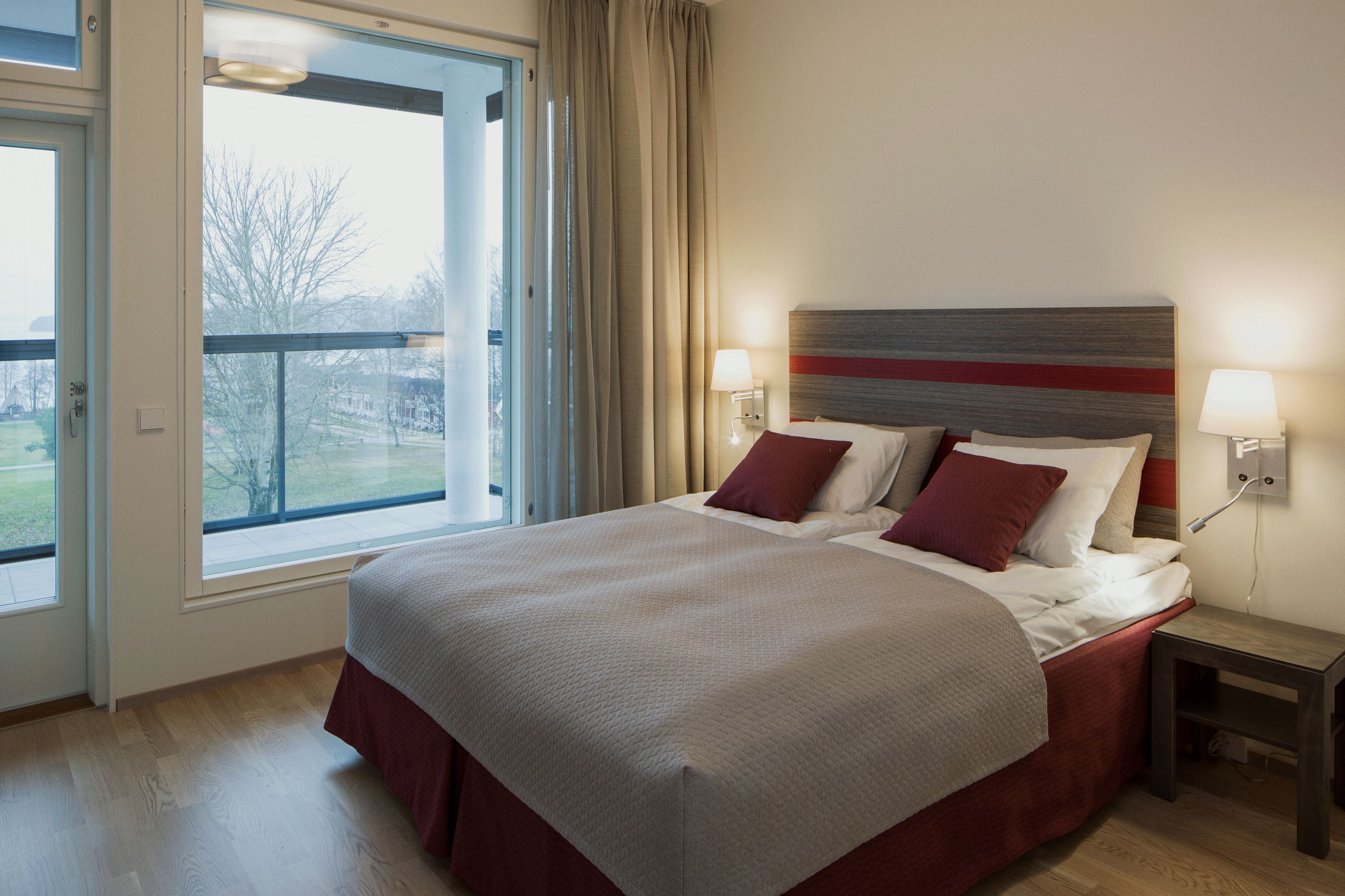 Una cama bien hecha con una decoración roja y blanca, una mesita de noche con lámpara y un ventanal con cortinas.