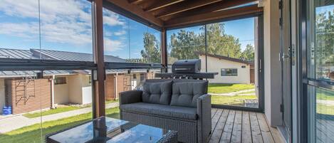 Un espacio al aire libre techado con una mesa de vidrio, mobiliario de mimbre, una parrilla y vistas a una zona residencial.
