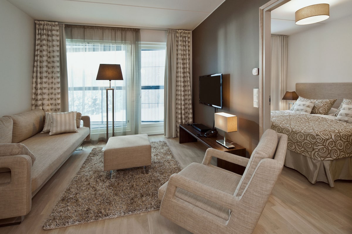 A modern hotel room with a sofa, a chair, a bed, a TV, and a desk with a lamp.