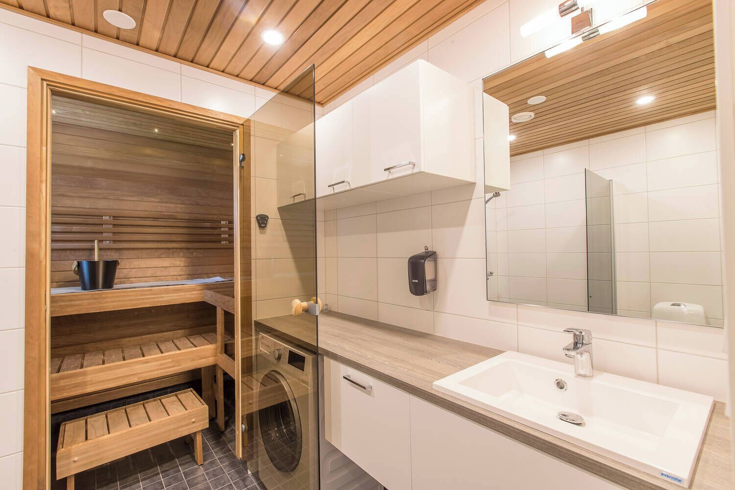 Un baño moderno con sauna de madera, lavadora en gabinete de vidrio y un espejo grande sobre el lavamanos.