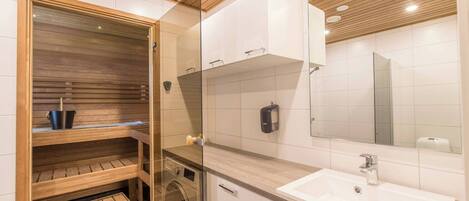 Un baño moderno con sauna de madera, lavadora en gabinete de vidrio y un espejo grande sobre el lavamanos.