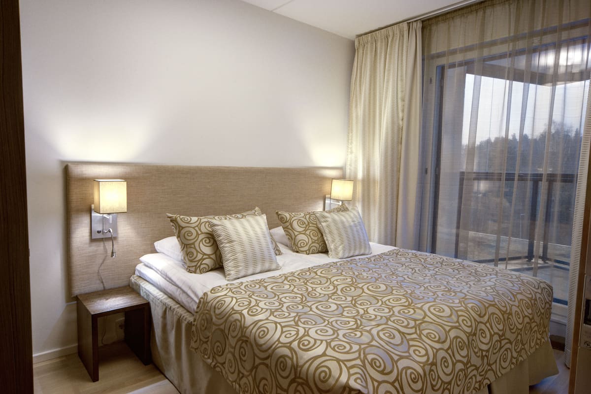 A hotel room with a large bed, two bedside lamps, a nightstand, and a view through a large window.