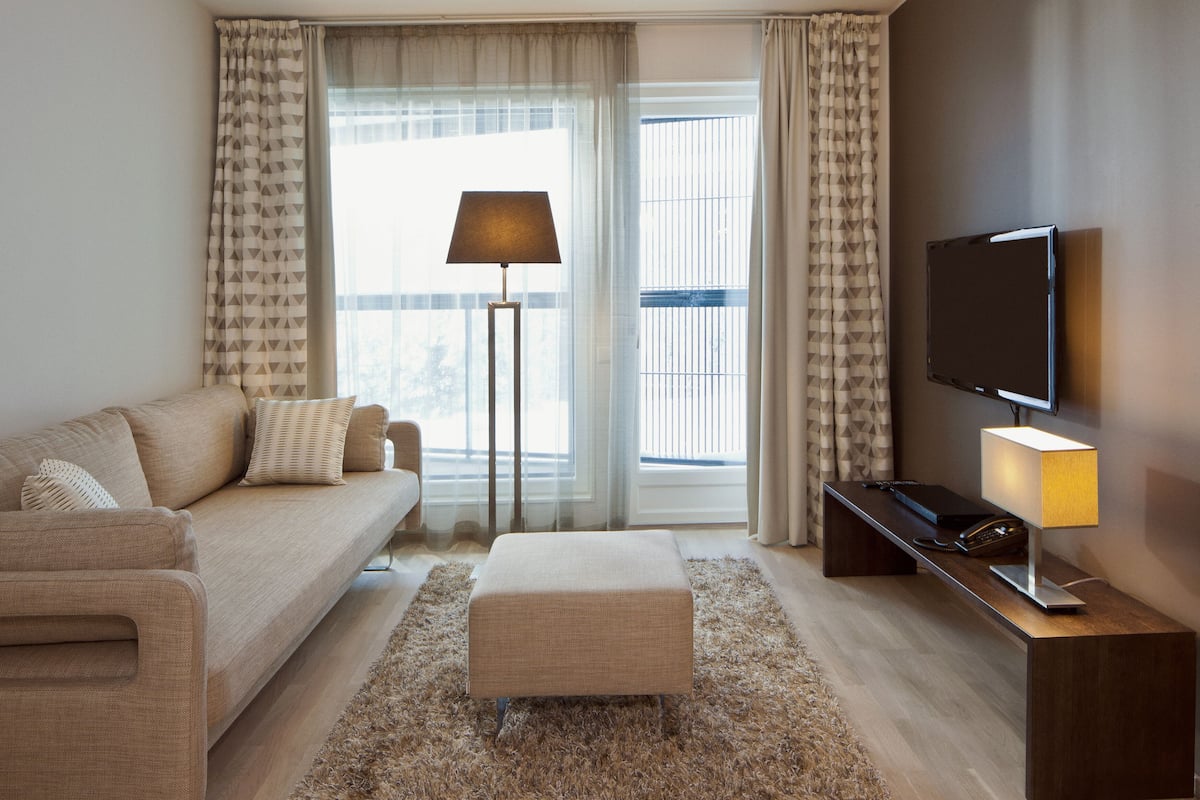 A modern living room with a sofa, ottoman, floor lamp, TV, and a view of the outdoors through large windows.