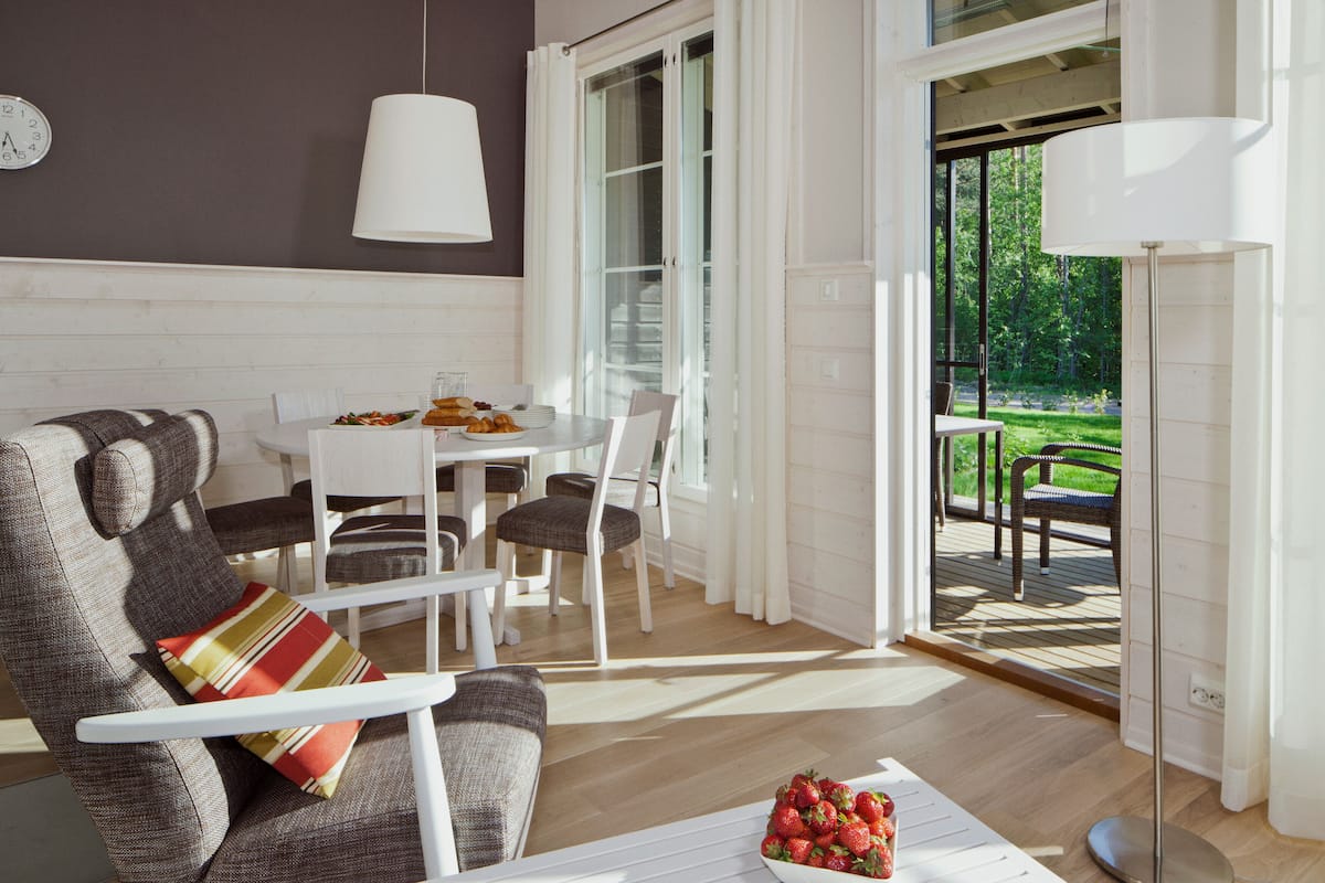 A modern living room with a sofa, dining table, and chairs. There is a view of a garden through the glass door.