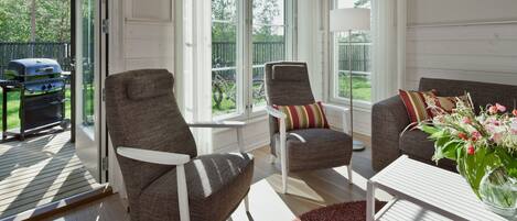 A room with a sliding glass door leading to a patio, two upholstered chairs, a white coffee table, and a floral arrangement.
