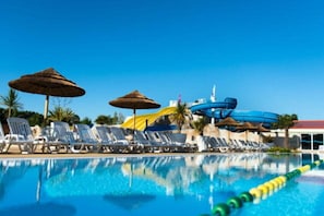 A swimming pool with lounge chairs and umbrellas, a water slide, and a building in the background.