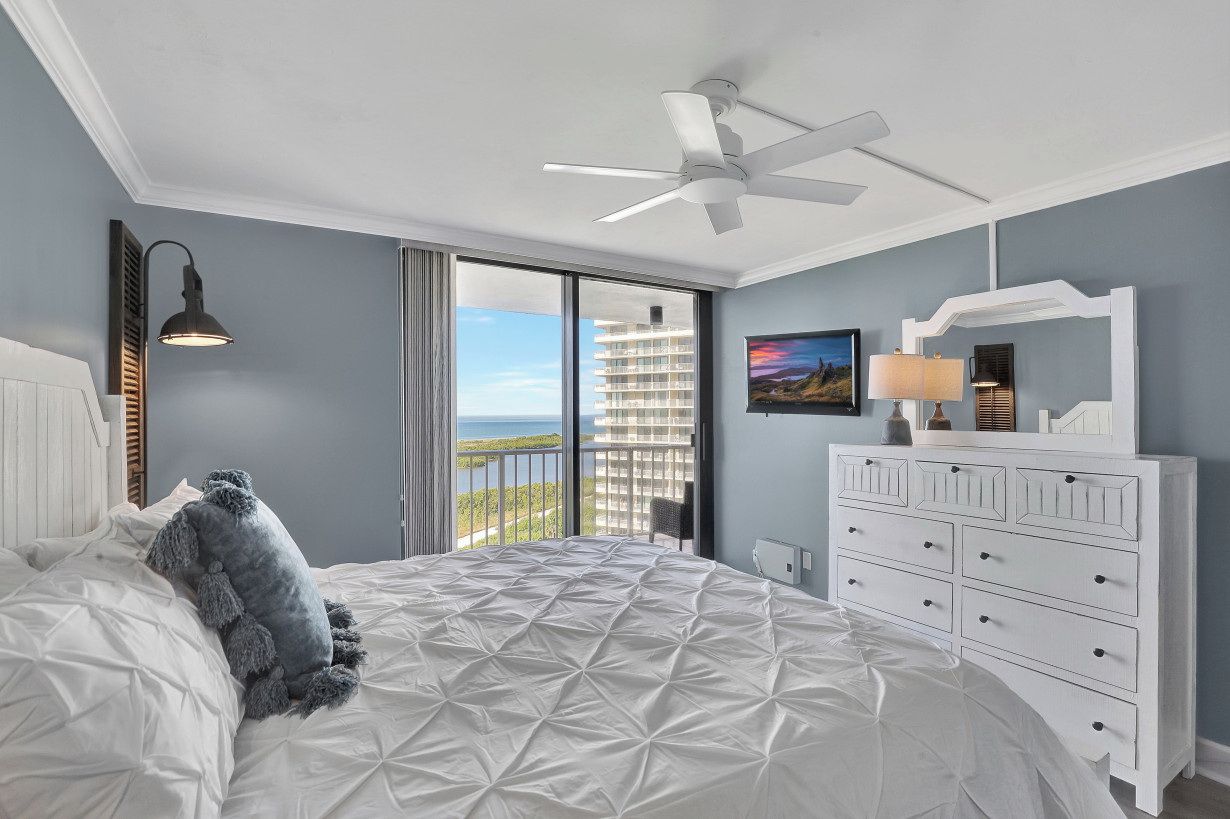 Une chambre avec un grand lit, une commode blanche, un ventilateur de plafond, une fenêtre donnant sur un immeuble et une table de chevet avec une lampe.