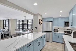 A modern kitchen with light blue cabinets, a large island, stainless steel appliances, and a dining area with a view of the city.